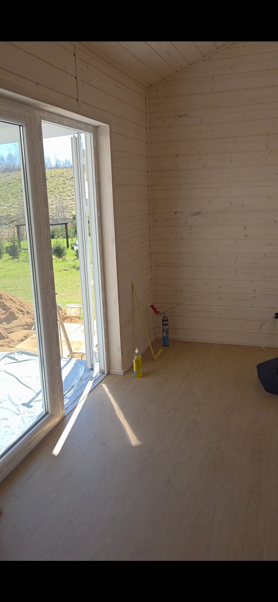 Wnętrze surowego domu z drewnianą boazerią, widoczne okno z widokiem na zewnątrz, butelka płynu, puszka pianki montażowej i miara zwinięta przy ścianie.