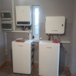 MARIUSZ KOWALCZYK CREON GROUP - Dwa białe magazyny energii Dyness w pomieszczeniu, z podłączonymi kablami, obok inwerter i skrzynki elektryczne. Widoczne okno i płytki na podłodze.