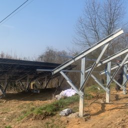 MARIUSZ KOWALCZYK CREON GROUP - Montaż konstrukcji wsporczej pod panele słoneczne na nierównym terenie. Częściowo zamontowane panele, widoczne narzędzia i pracownik w tle. Słoneczny dzień.