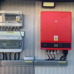 MARIUSZ KOWALCZYK CREON GROUP - Czerwony falownik GoodWe i rozdzielnice Noark na metalowej ścianie. Widoczne kable, oznaczenia i lampa LED. Instalacja fotowoltaiczna w szczegółach.