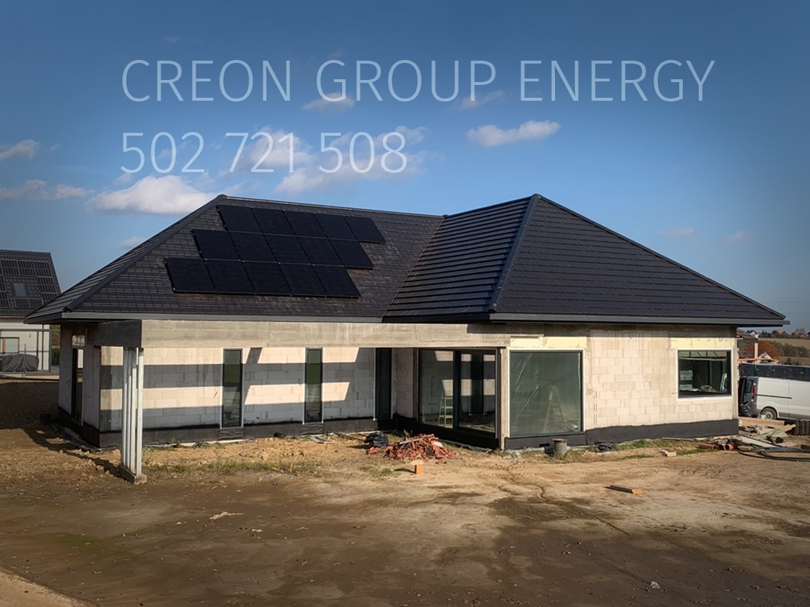 Dom jednorodzinny w trakcie budowy z zamontowanymi panelami fotowoltaicznymi na dachu. Widoczne bloczki betonowe i duże okna. Na górze logo firmy i numer telefonu.