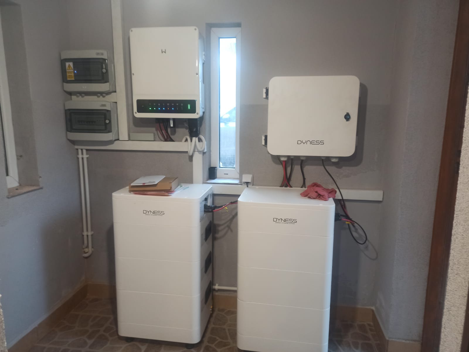 Dwa białe magazyny energii Dyness w pomieszczeniu, z podłączonymi kablami, obok inwerter i skrzynki elektryczne. Widoczne okno i płytki na podłodze.