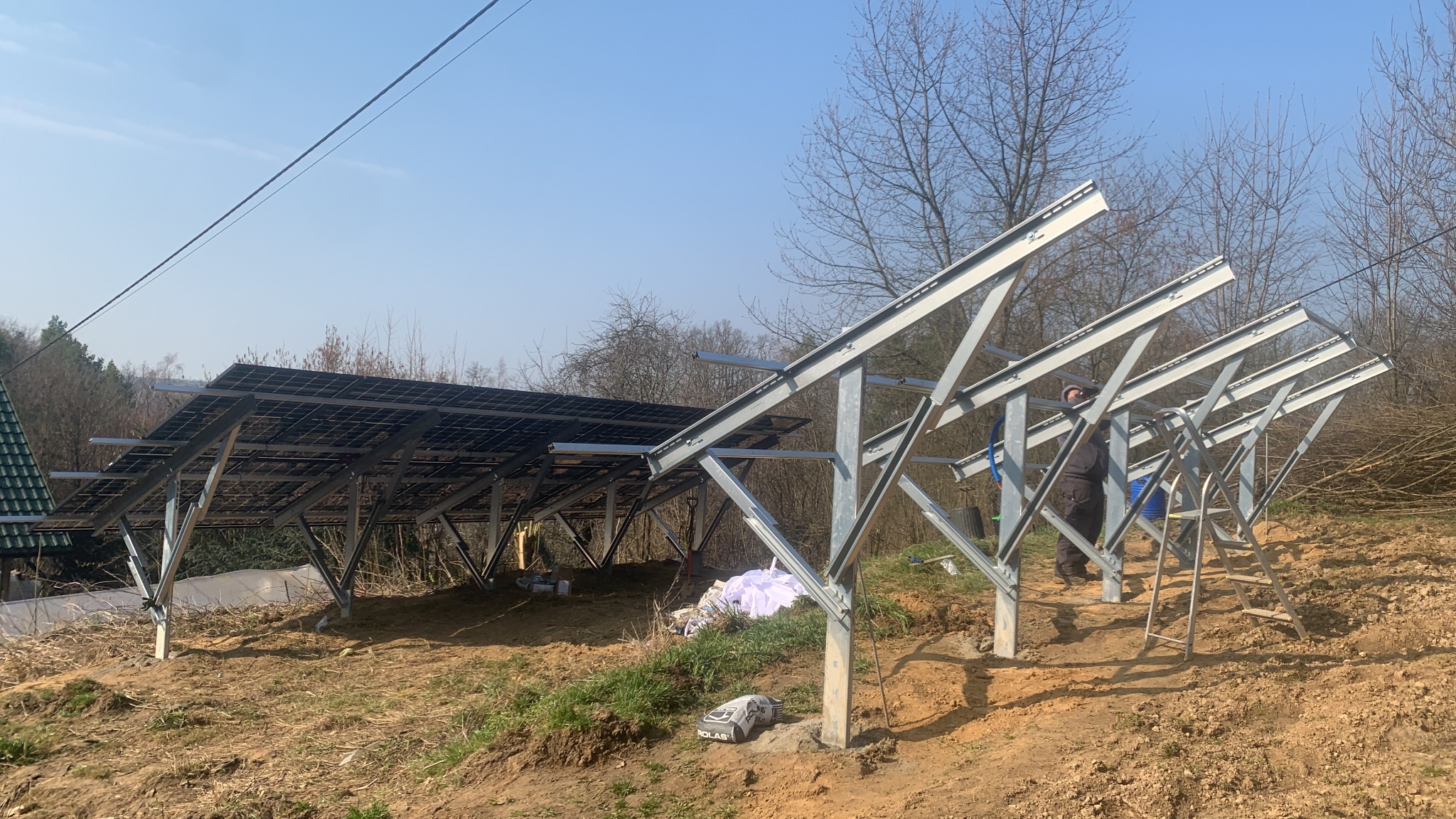 Montaż konstrukcji wsporczej pod panele słoneczne na nierównym terenie. Częściowo zamontowane panele, widoczne narzędzia i pracownik w tle. Słoneczny dzień.