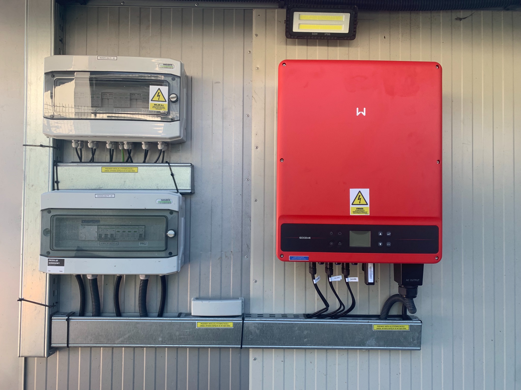 Czerwony falownik GoodWe i rozdzielnice Noark na metalowej ścianie. Widoczne kable, oznaczenia i lampa LED. Instalacja fotowoltaiczna w szczegółach.