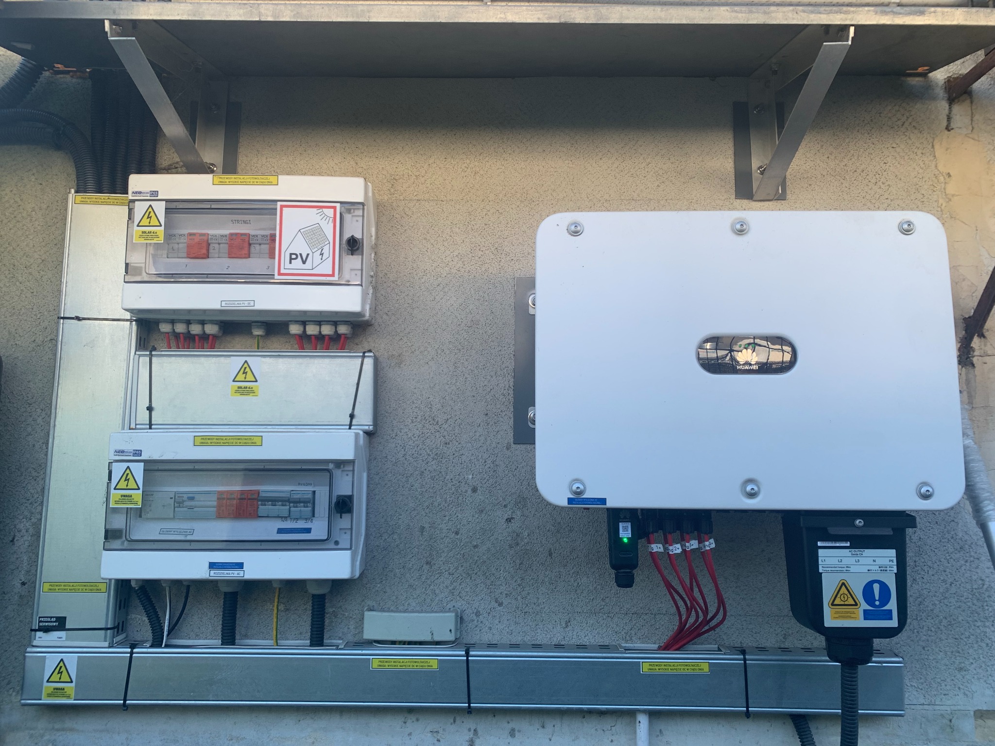Instalacja fotowoltaiczna: rozdzielnice i inwerter Huawei zamontowane na szarej ścianie. Widoczne kable i oznaczenia ostrzegawcze.