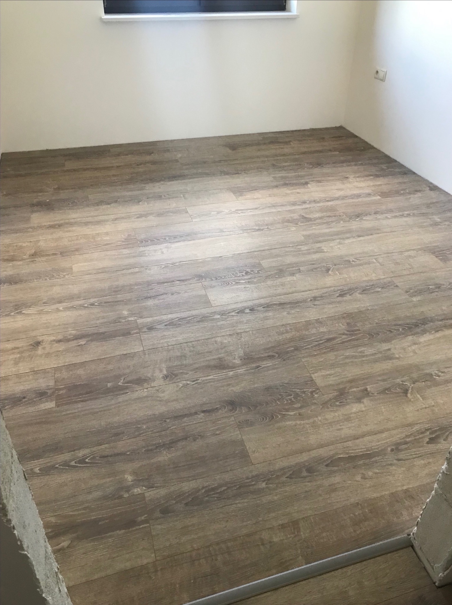 Świeżo ułożona podłoga z paneli imitujących drewno w pustym pokoju z białymi ścianami i oknem u góry, widoczny fragment progu.