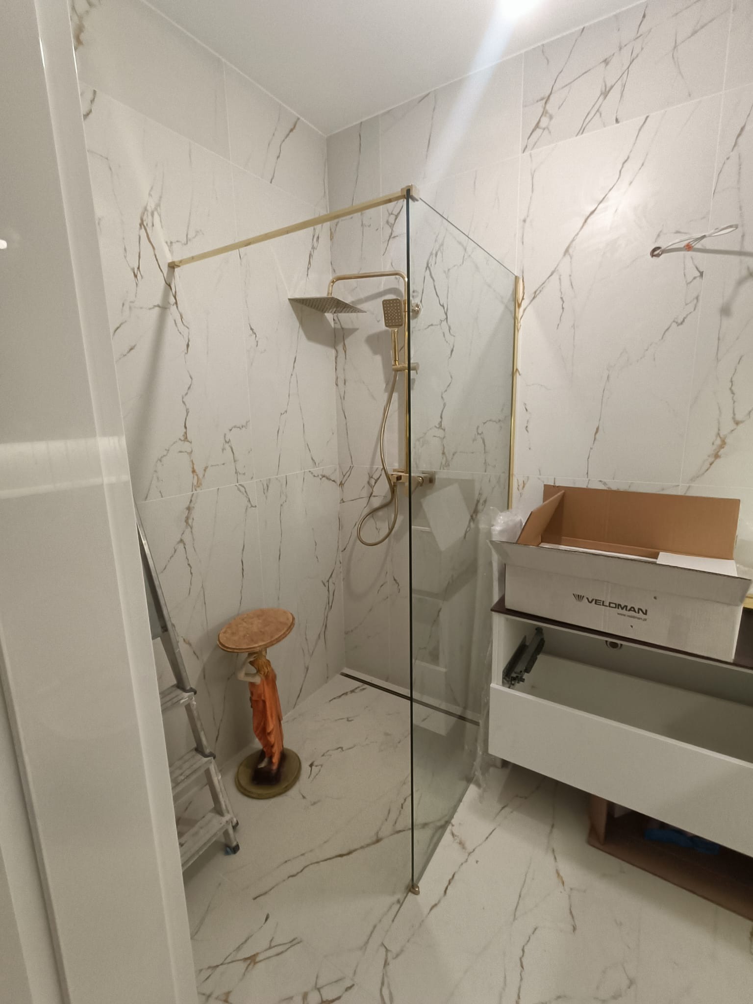 Szklana kabina prysznicowa z złotymi elementami, zamontowana w łazience wyłożonej marmurowymi płytkami. Widoczna drabina, podest z figurką i otwarta szafka z pudełkiem Veloman.