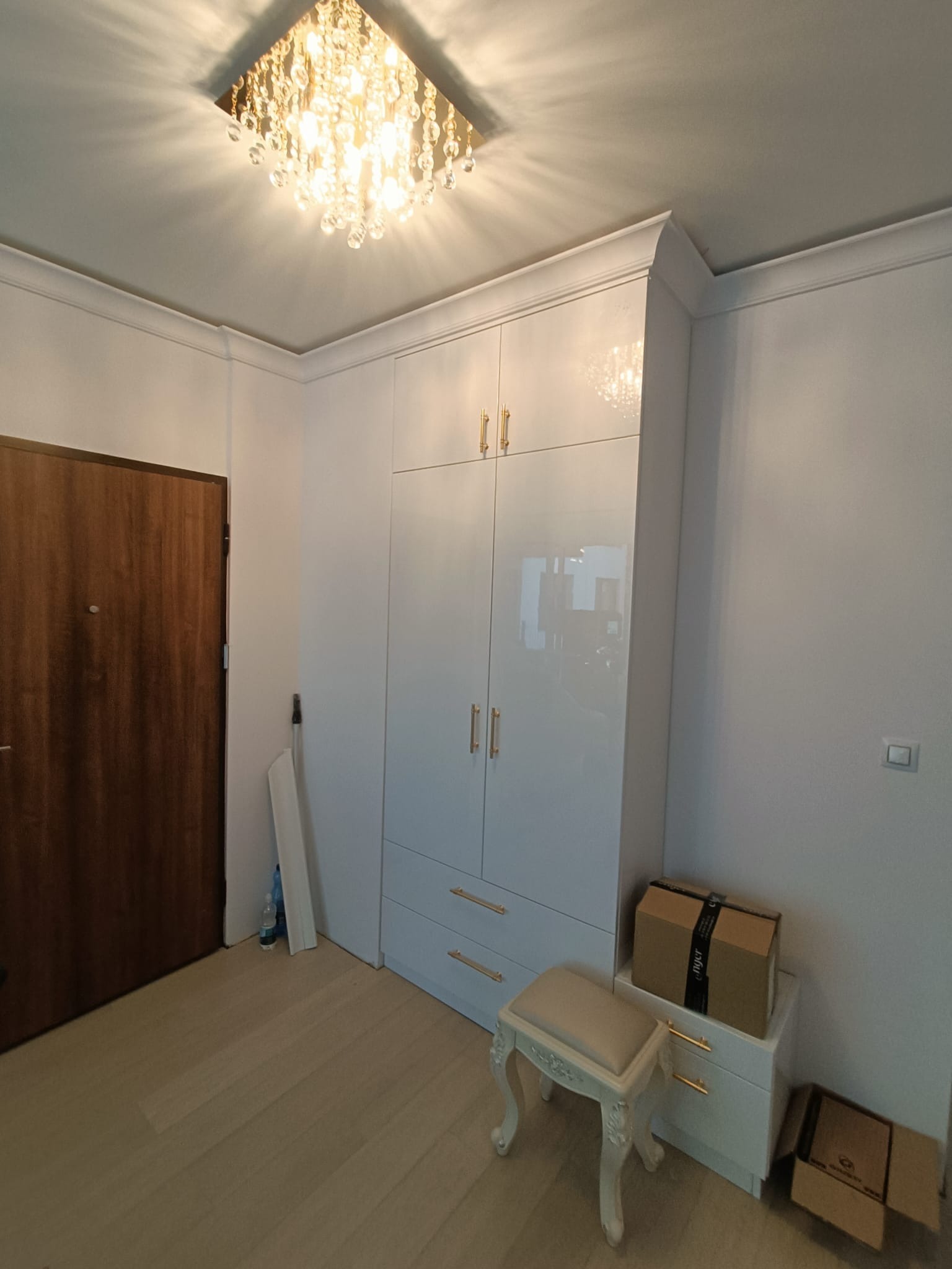 Biała, lakierowana szafa wnękowa z złotymi uchwytami, dopasowana do sufitu z ozdobnym, kryształowym żyrandolem; obok drzwi wejściowe i pufa w stylu ludwikowskim.