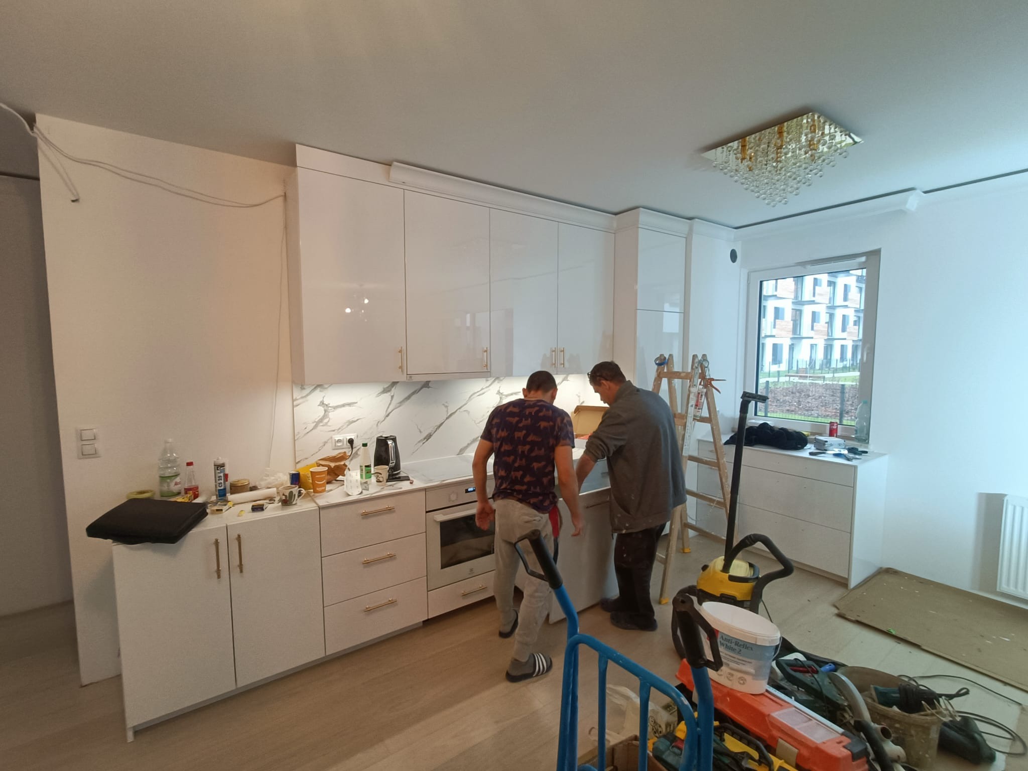 Montaż białej, lakierowanej kuchni z marmurowym blatem i złotymi uchwytami; dwaj monterzy w trakcie pracy, widoczny wózek z narzędziami i drabina.