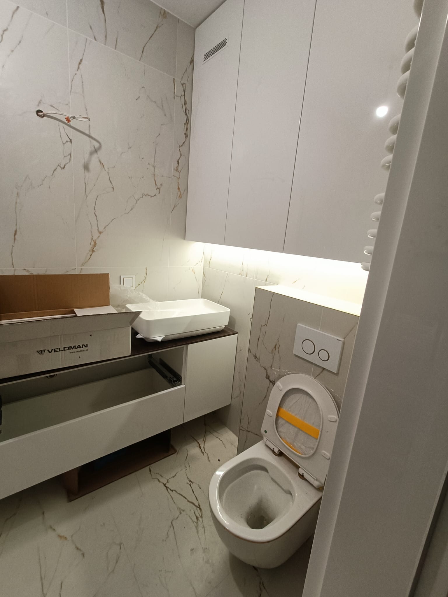 Łazienka w trakcie montażu: umywalka nablatowa w kartonie marki Veloman, otwarta szafka pod umywalkę, wisząca toaleta z żółtą folią zabezpieczającą na desce, płytki imitujące marmur na ścianach.