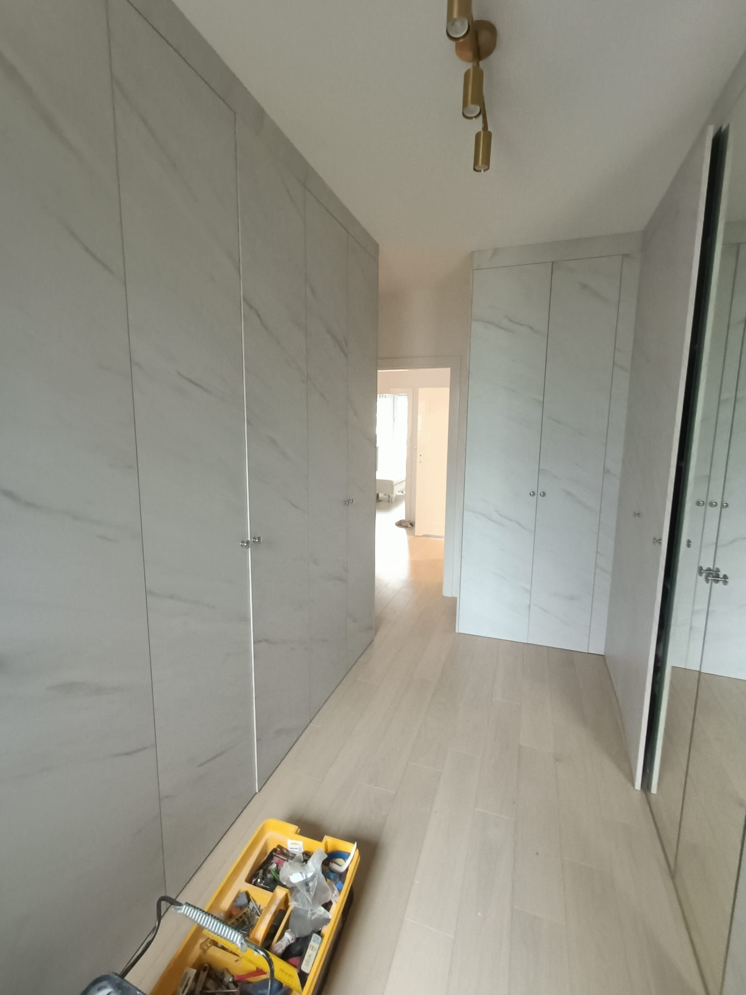 Długi korytarz z zabudową szaf wnękowych z frontami imitującymi marmur, widoczna żółta skrzynka z narzędziami, sufitowa lampa w kolorze starego złota.