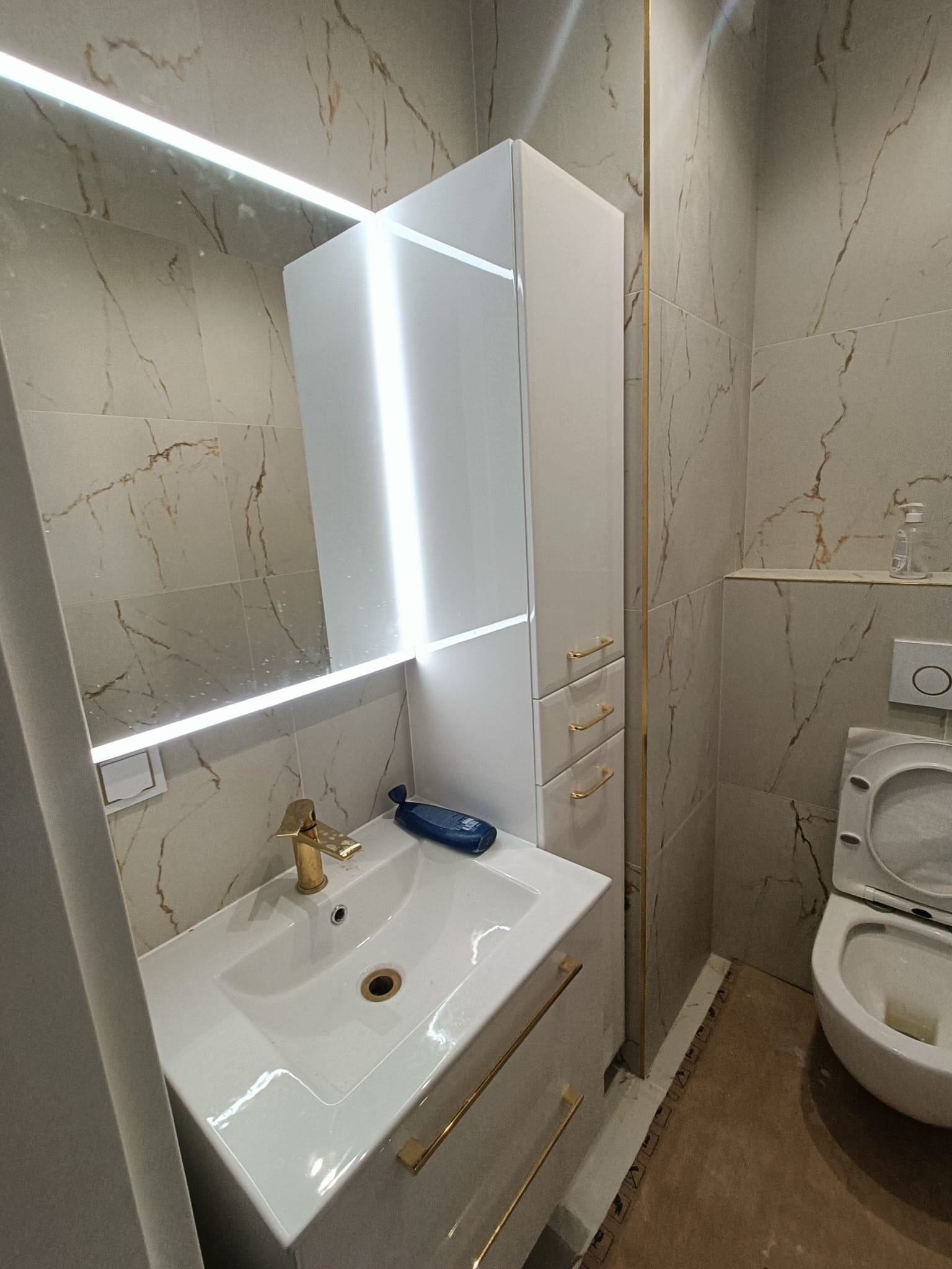 Nowoczesna łazienka z marmurowymi płytkami, podświetlanym lustrem, złotą armaturą i białymi meblami łazienkowymi z złotymi uchwytami. Widoczny fragment toalety i brązowa podłoga.