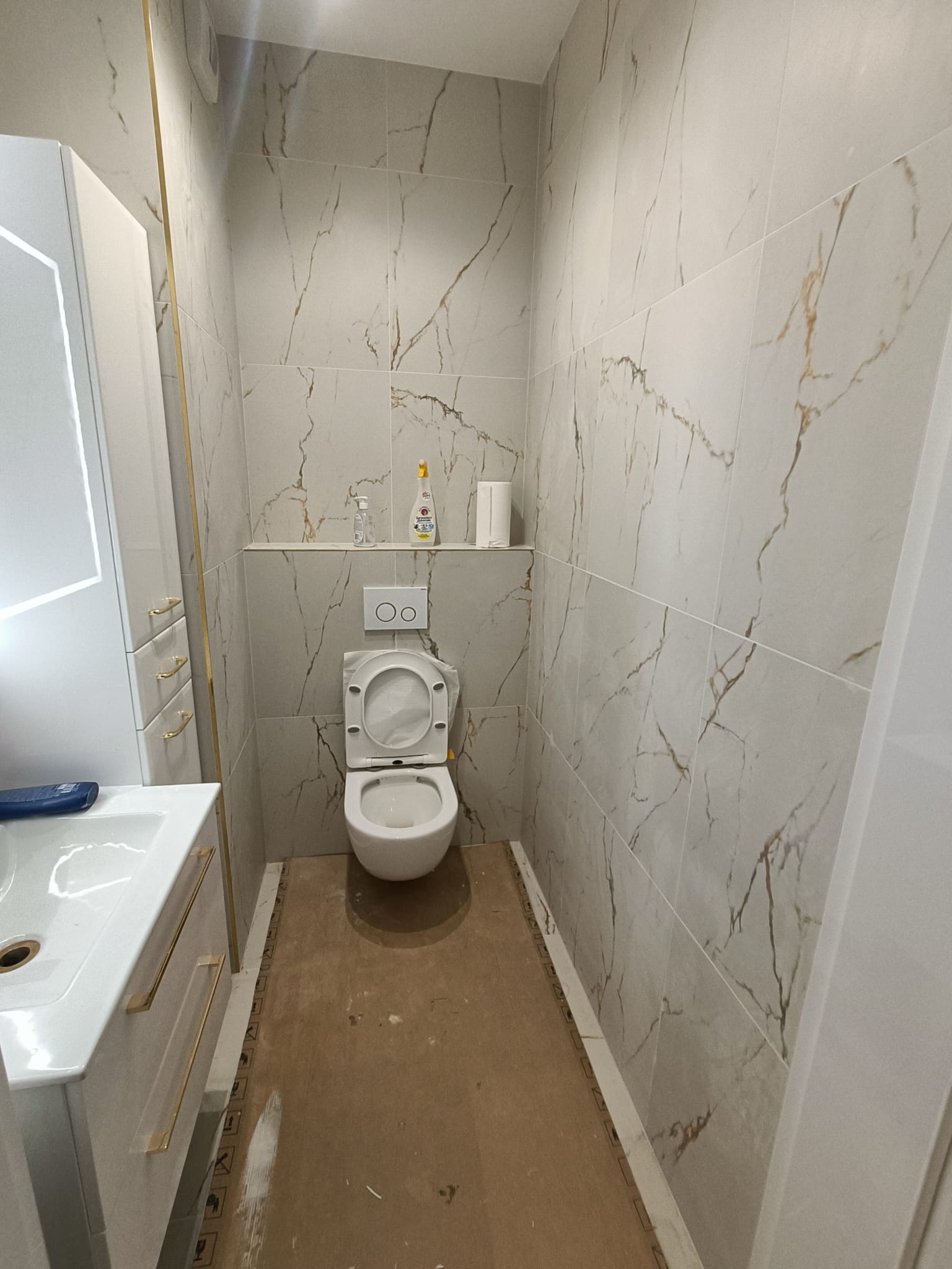Mała łazienka w trakcie prac wykończeniowych: białe marmurowe płytki na ścianach, biała toaleta z otwartą klapą, biała szafka z umywalką i złotymi uchwytami, podłoga zabezpieczona kartonem.