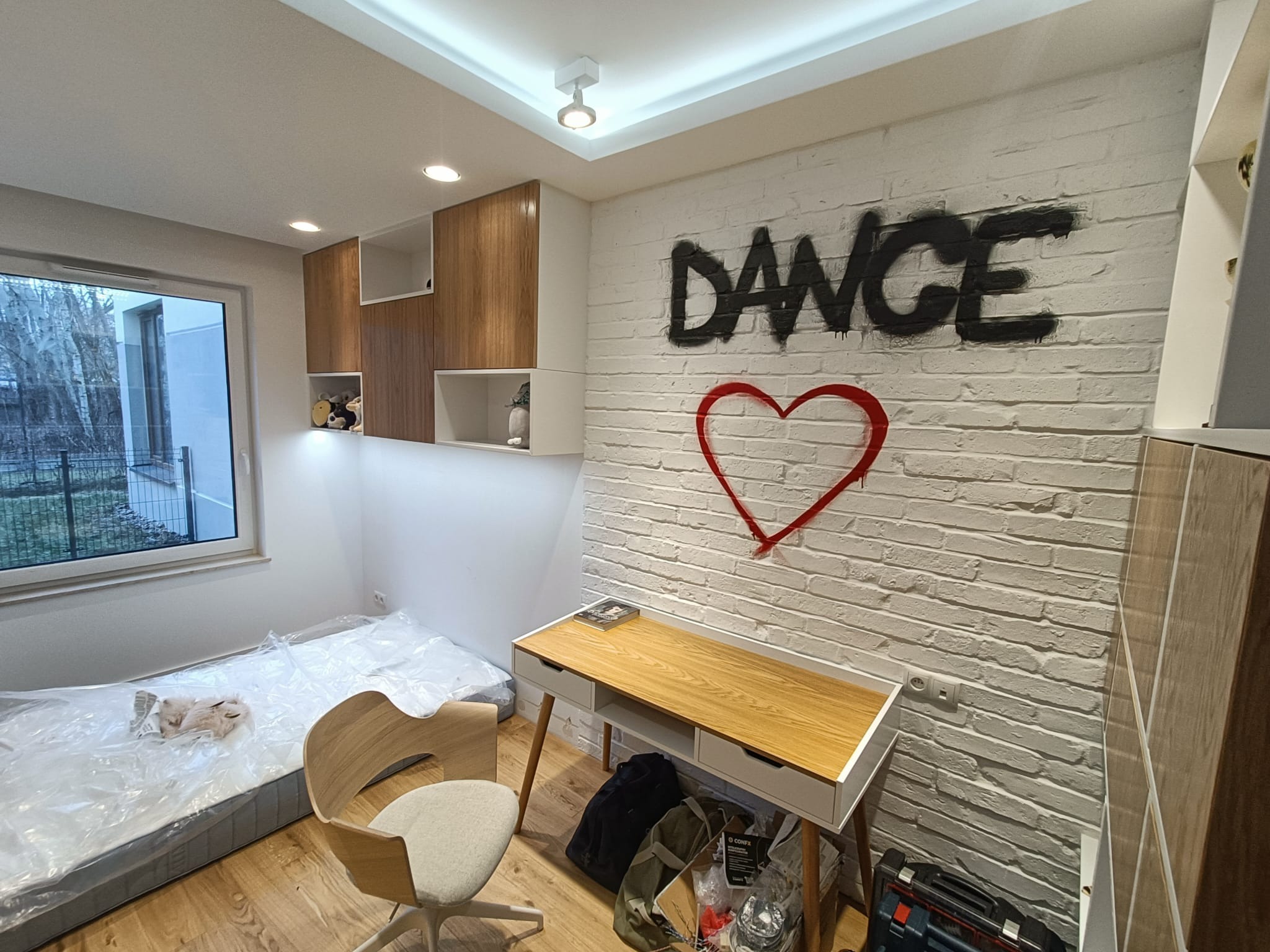 Pokój młodzieżowy z białej cegły z napisem DANCE i sercem, biurko z drewnianym blatem, krzesło na kółkach i materac w folii ochronnej.
