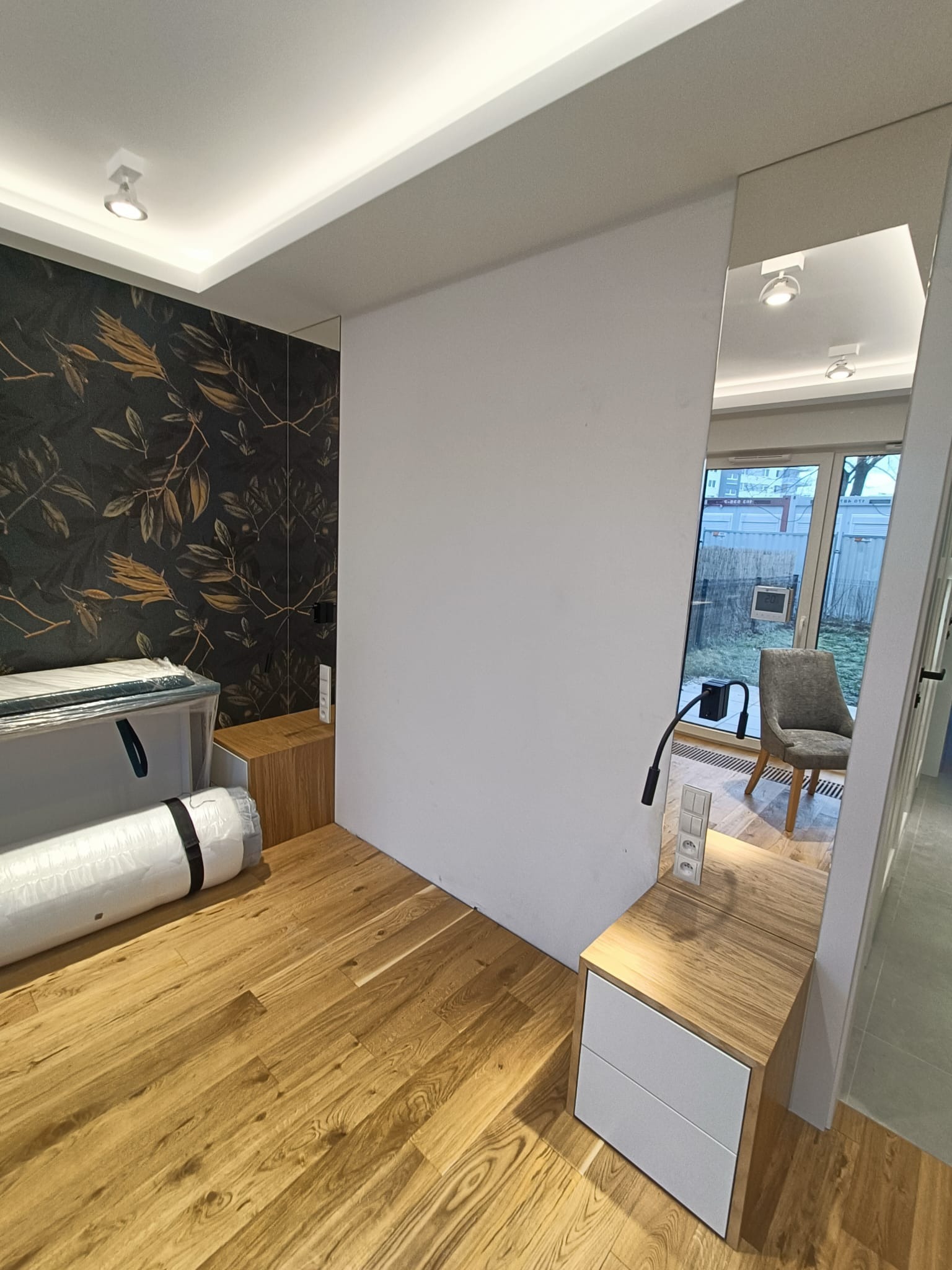 Pokój w trakcie aranżacji: drewniana podłoga, szafka nocna z białymi szufladami i gniazdkami, tapeta w ciemne liście, zwinięty materac w folii, lustro odbijające krzesło i okno.