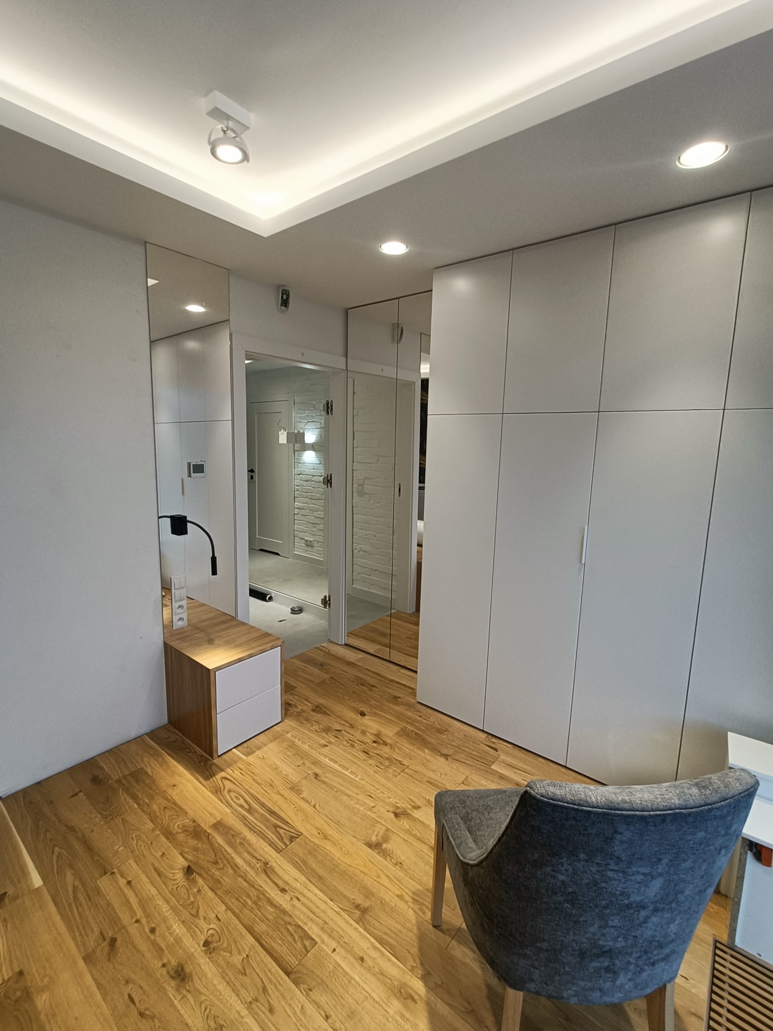Nowoczesna garderoba z lustrzanymi drzwiami, drewnianą podłogą i szarym fotelem, widok na korytarz z ceglaną ścianą.
