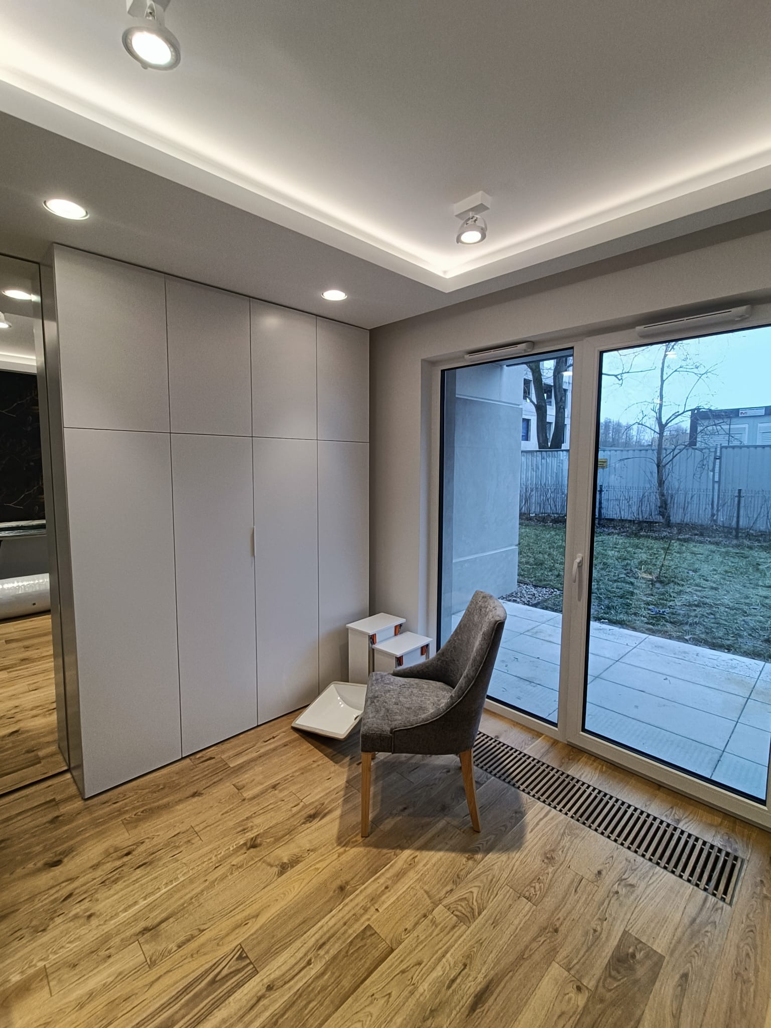 Nowoczesny pokój z dużą szafą w zabudowie, szarym fotelem i przesuwnymi drzwiami balkonowymi z widokiem na ogród.