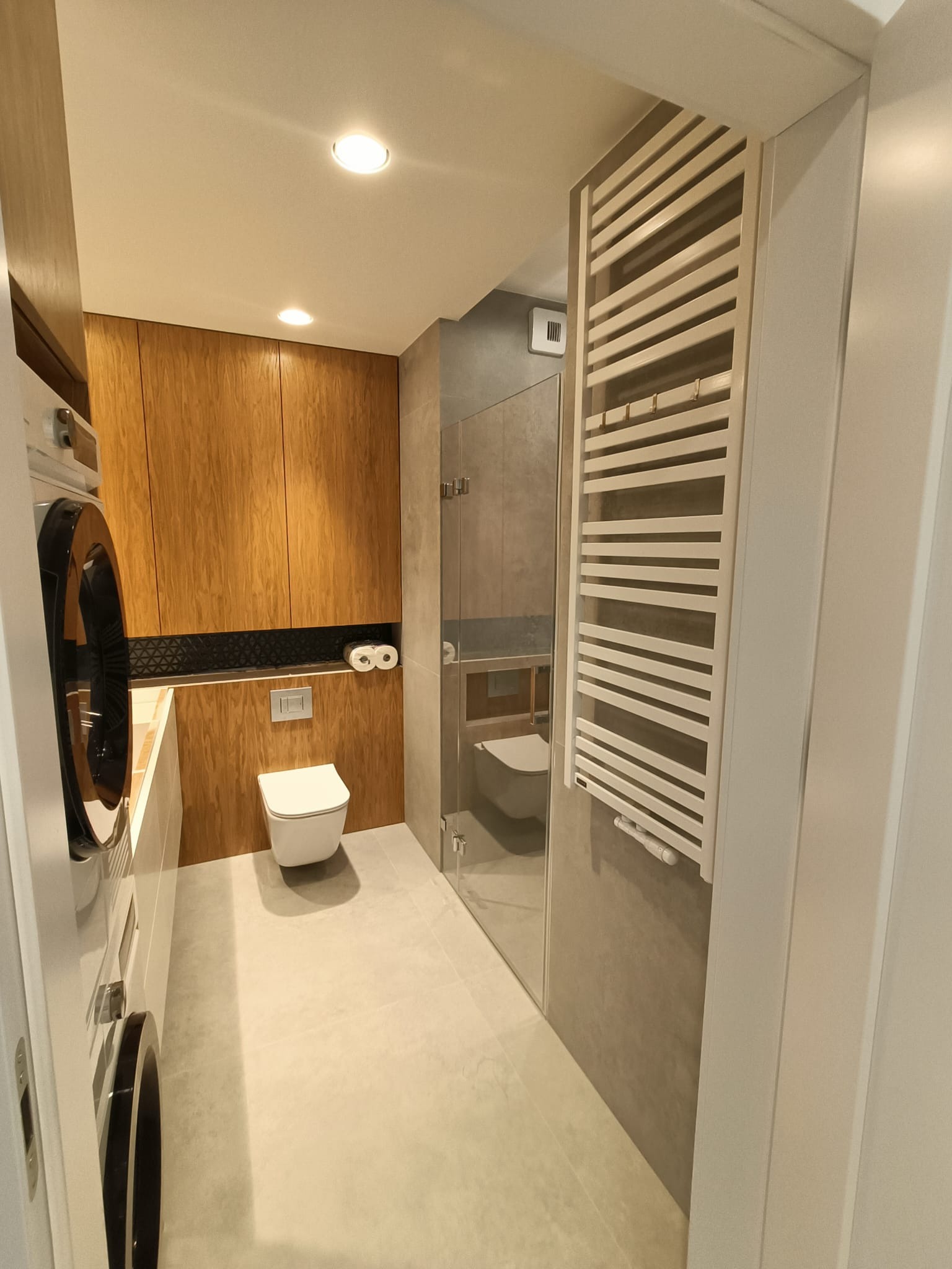 Nowoczesna łazienka z drewnianymi szafkami, pralką z czarnym bębnem po lewej stronie, białą toaletą podwieszaną i grzejnikiem drabinkowym po prawej, widok zza otwartych drzwi.