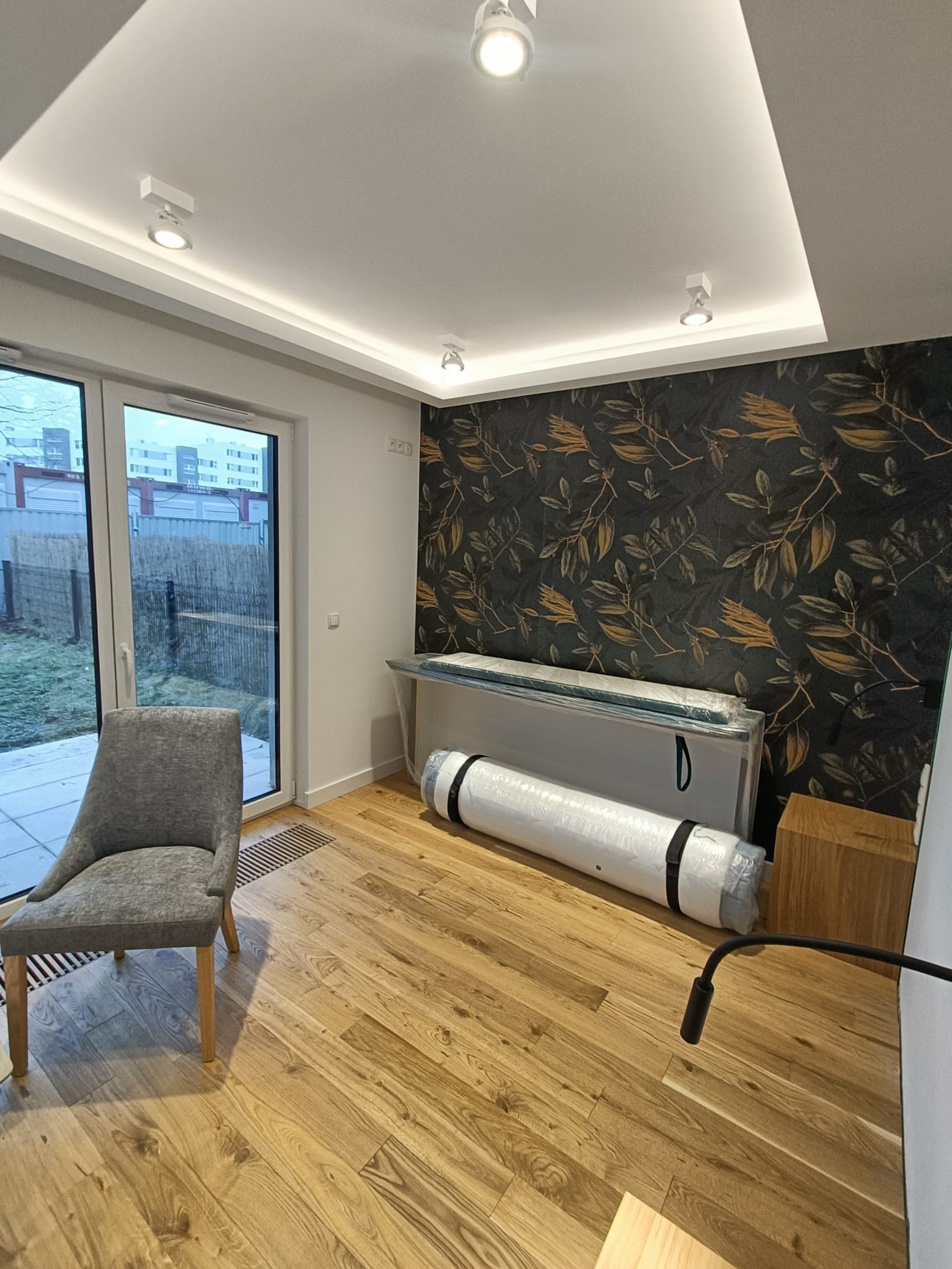 Pokój z drewnianą podłogą i tapetą w liście, z szarym krzesłem i meblami w folii ochronnej, przygotowany do montażu.