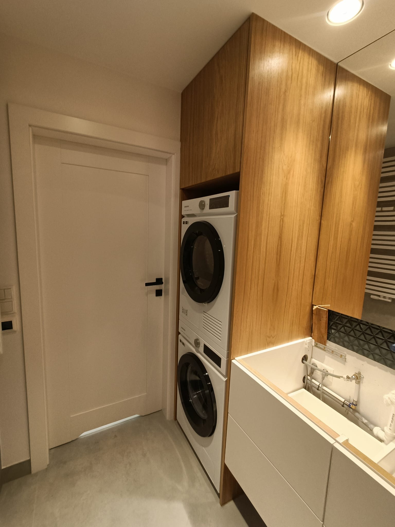 Wnętrze łazienki z pralką i suszarką w zabudowie z drewnianymi frontami, biała umywalka z widoczną instalacją hydrauliczną, białe drzwi z czarną klamką.