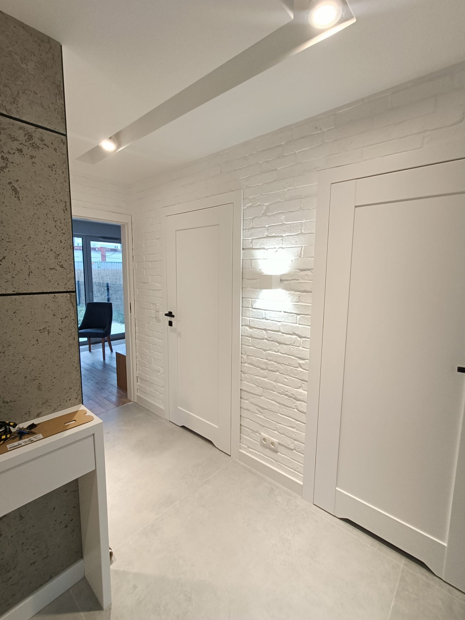 Jasne wnętrze przedpokoju z białymi drzwiami, ścianą z białej cegły i szarą podłogą. Widoczne oświetlenie sufitowe punktowe i dekoracyjna lampa ścienna. W tle widoczny pokój z krzesłem.