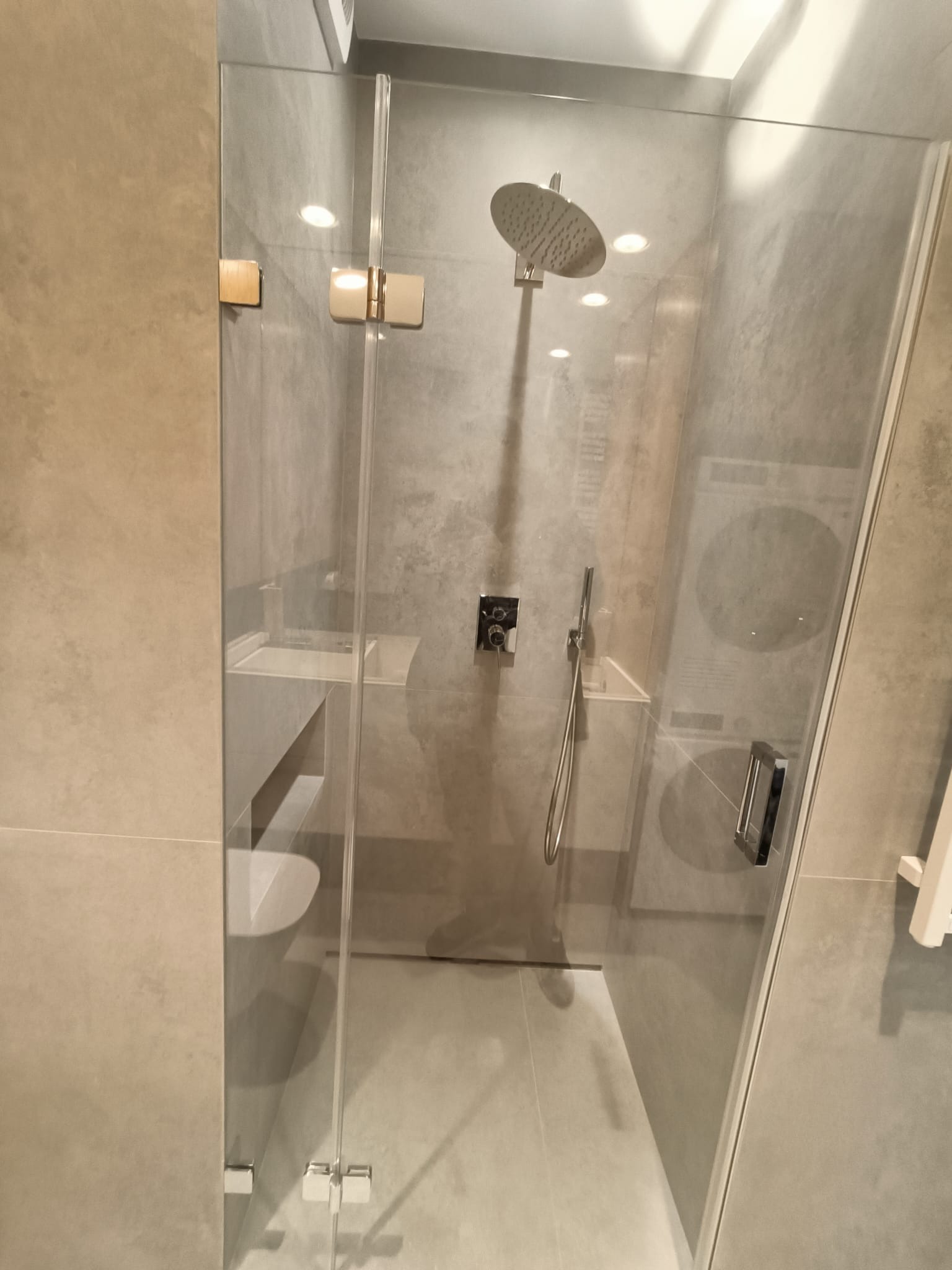 Szklana kabina prysznicowa z deszczownicą i baterią termostatyczną, umywalka nablatowa z szafką w szarym kolorze, widoczna pralka w tle, minimalistyczny design łazienki.