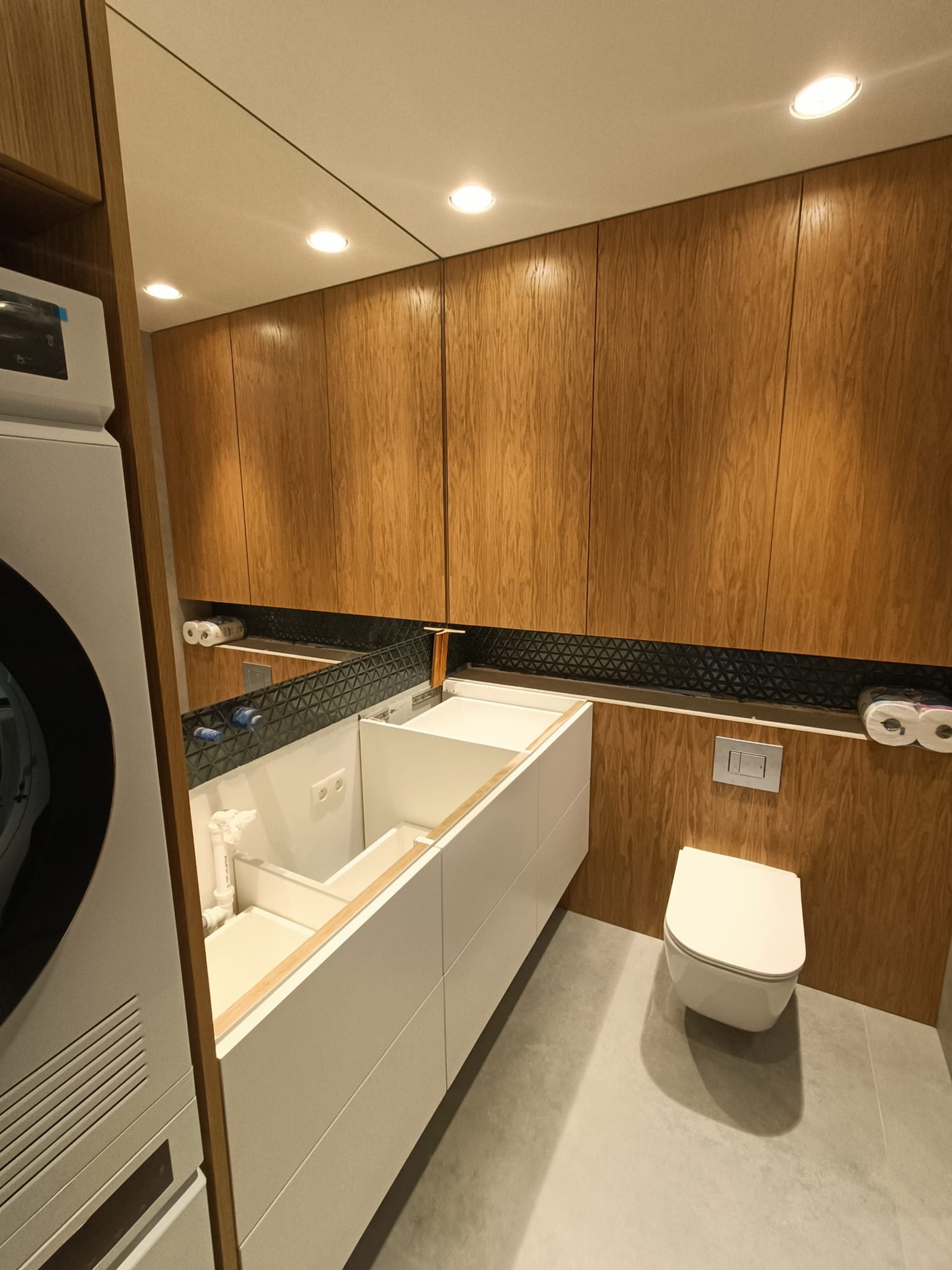 Nowoczesna łazienka w trakcie wykańczania: biała umywalka w zabudowie, wisząca toaleta, drewniane szafki z czarną mozaiką, pralka z boku.