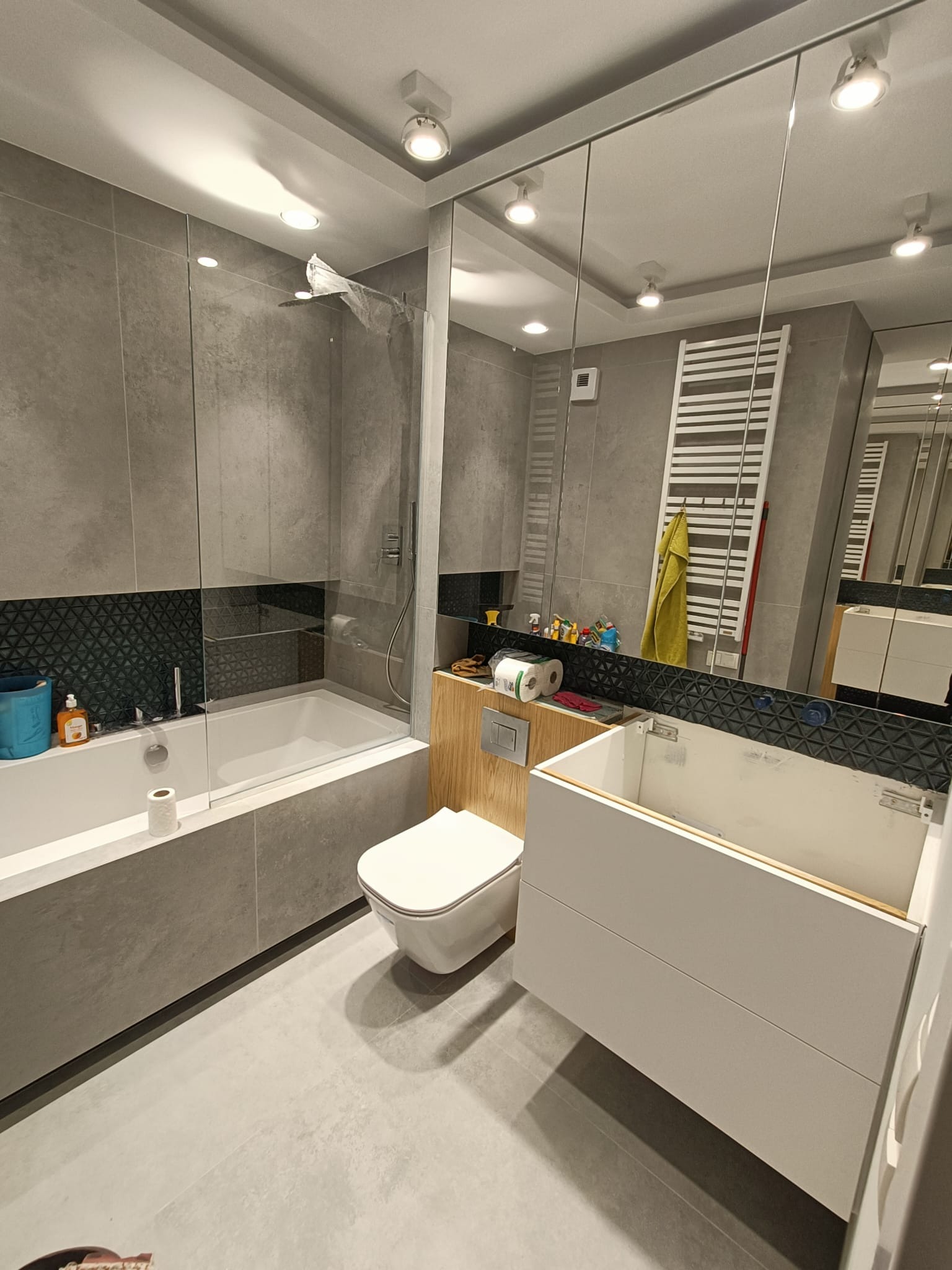 Nowoczesna łazienka w szarościach z elementami drewna, widoczna wanna z parawanem, toaleta i szafka pod umywalkę w trakcie montażu, lustro odbija grzejnik drabinkowy i akcesoria.