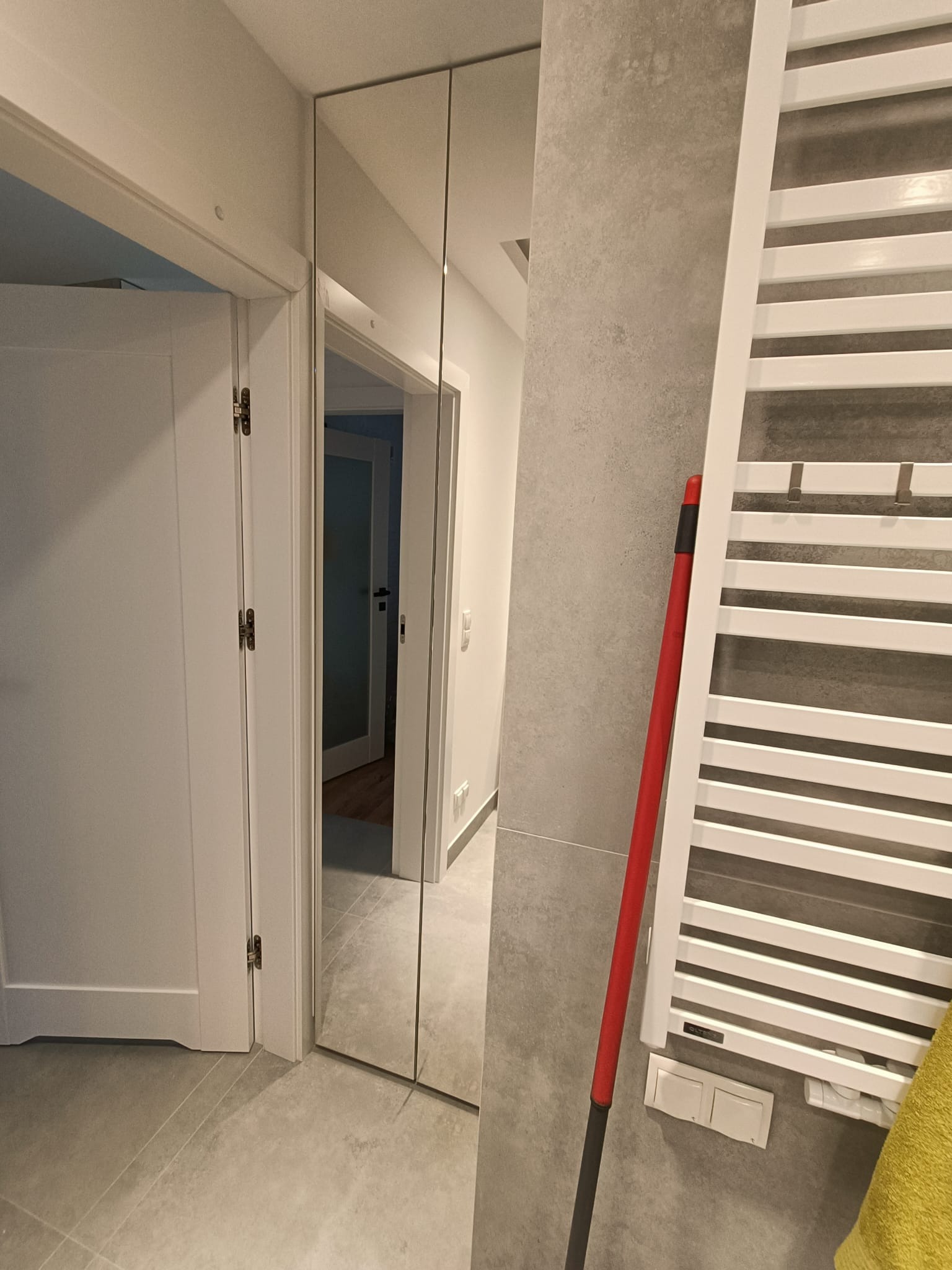 Szare płytki podłogowe i ścienne w korytarzu, lustro w zabudowie, biały grzejnik drabinkowy z żółtym ręcznikiem i czerwonym mopem, widok na otwarte drzwi do pokoju.