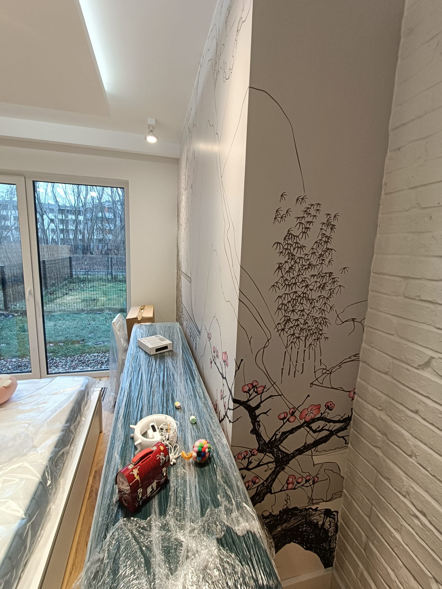 Pokój w trakcie prac wykończeniowych: ściana z motywem drzewa wiśni, zabezpieczone folią meble, zabawki i lunchbox na blacie, widok na ogród przez okno.