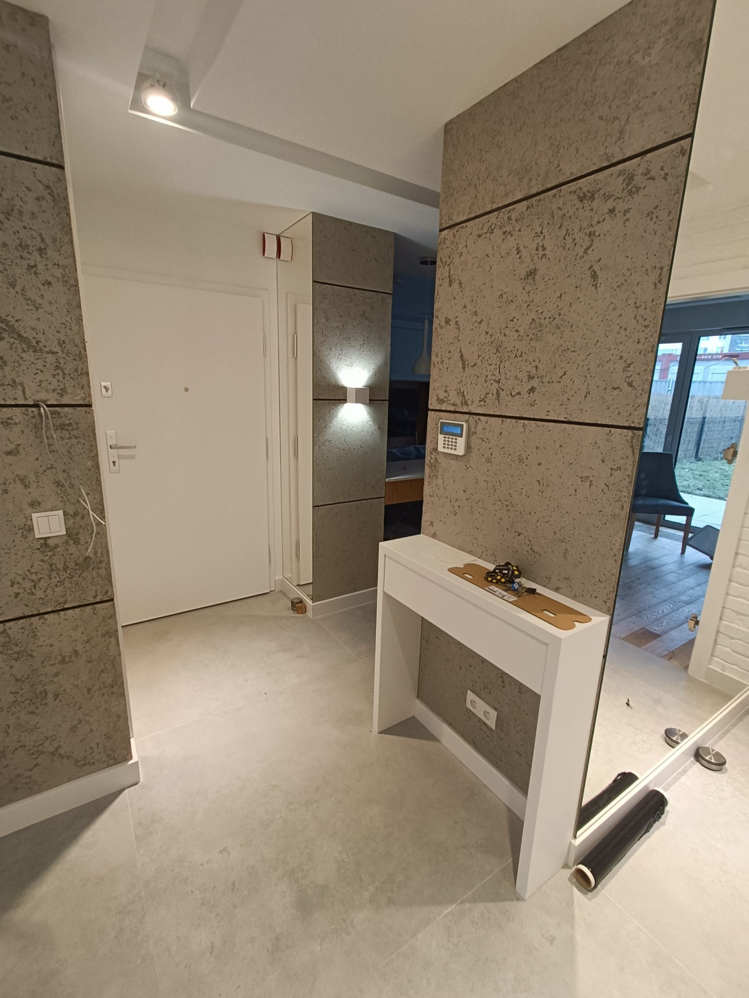 Nowoczesny przedpokój z panelami dekoracyjnymi o strukturze betonu, białą konsolą i dużym lustrem; widoczne detale wykończeniowe, takie jak oświetlenie i alarm.