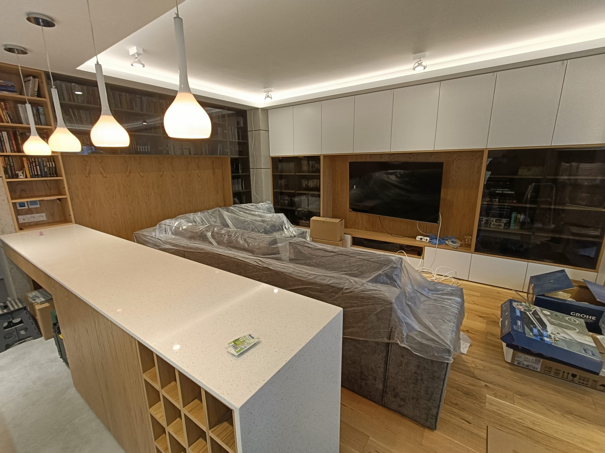 Salon w trakcie remontu: sofa zabezpieczona folią, widoczny blat z szafkami w zabudowie, regały z książkami, kartony z armaturą Grohe na podłodze.