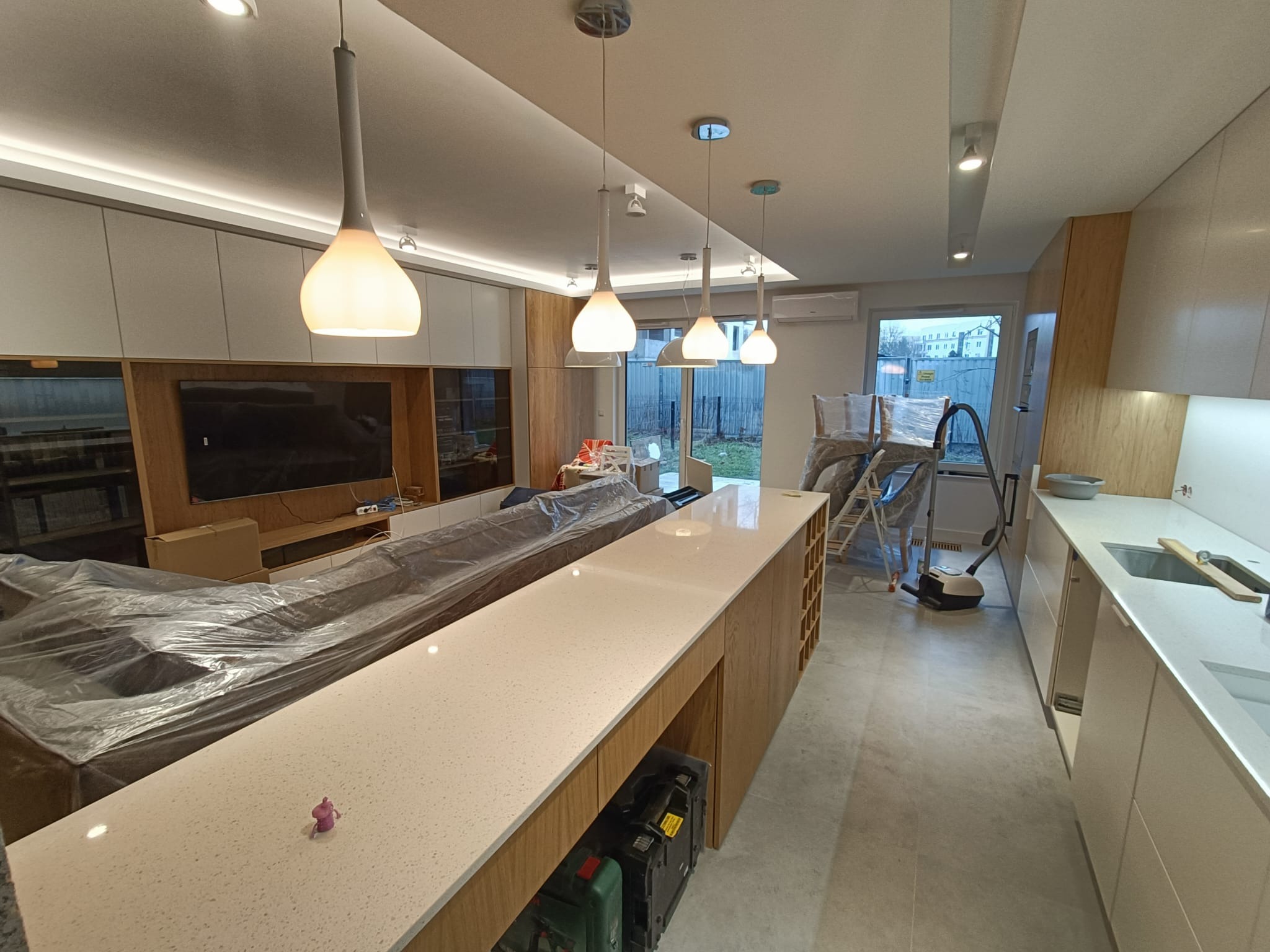 Jasne, nowoczesne wnętrze w trakcie prac wykończeniowych: widoczna wyspa kuchenna z blatem z konglomeratu, sofa zabezpieczona folią, drabina malarska i odkurzacz w tle, wiszące lampy z mlecznego...