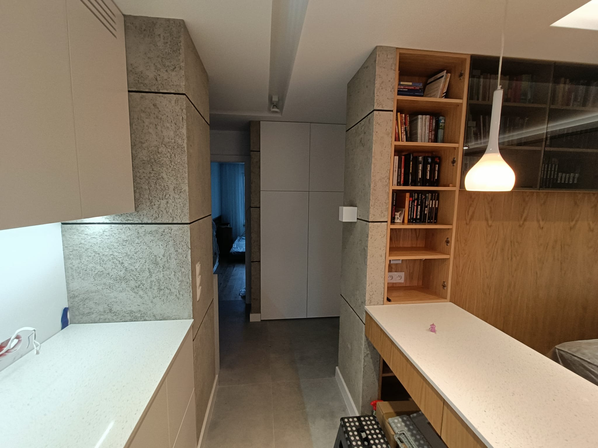 Wnętrze mieszkania z zabudową kuchenną w odcieniach szarości i drewna, oświetlone wiszącą lampą z mlecznego szkła, widok na korytarz z szarymi płytkami podłogowymi i szafą w zabudowie.