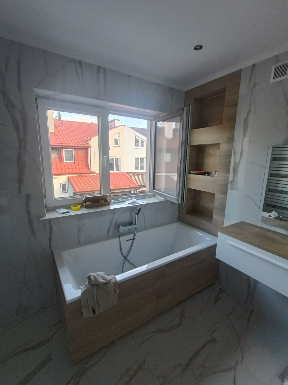 Nowoczesna łazienka z wanną obudowaną panelami drewnopodobnymi, marmurowe płytki na ścianach i podłodze, okno z widokiem na czerwone dachy budynków, wnęki w ścianie z półkami.