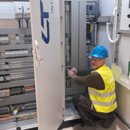 SIPRIX SPÓŁKA Z OGRANICZONĄ ODPOWIEDZIALNOŚCIĄ - Elektryk w niebieskim kasku i żółtej kamizelce, kuca przed otwartą szafą sterowniczą, podłączając przewody. Widoczna rozbudowana instalacja elektryczna wewnątrz szafy i na ścianie obok.