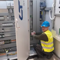 Elektryk w niebieskim kasku i żółtej kamizelce, kuca przed otwartą szafą sterowniczą, podłączając przewody. Widoczna rozbudowana instalacja elektryczna wewnątrz szafy i na ścianie obok.