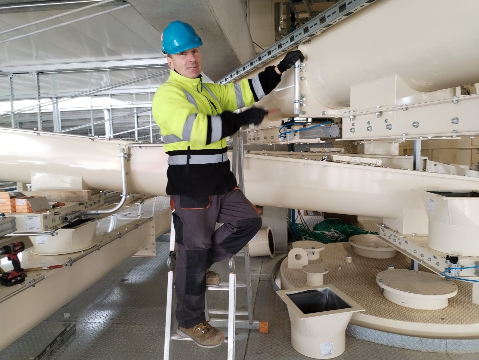 Elektryk w kasku i odblaskowej kurtce na drabinie podczas montażu instalacji w hali przemysłowej, z widocznym wiertarko-wkrętakiem obok.