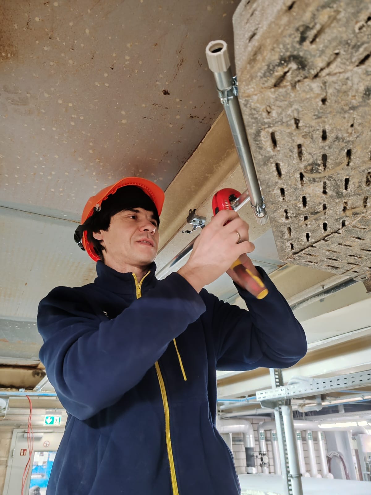 Elektryk w pomarańczowym kasku montuje metalową rurę do perforowanej belki pod sufitem, używając czerwono-żółtego narzędzia; widoczne instalacje rurowe w tle.