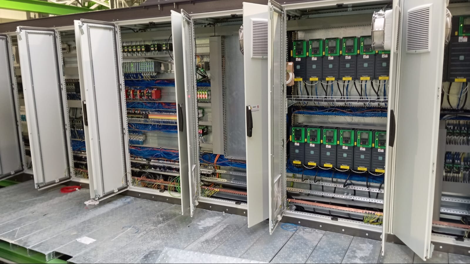 Otwarty rozdzielnica elektryczna z widocznymi przewodami, zabezpieczeniami i sterownikami na metalowej podłodze.