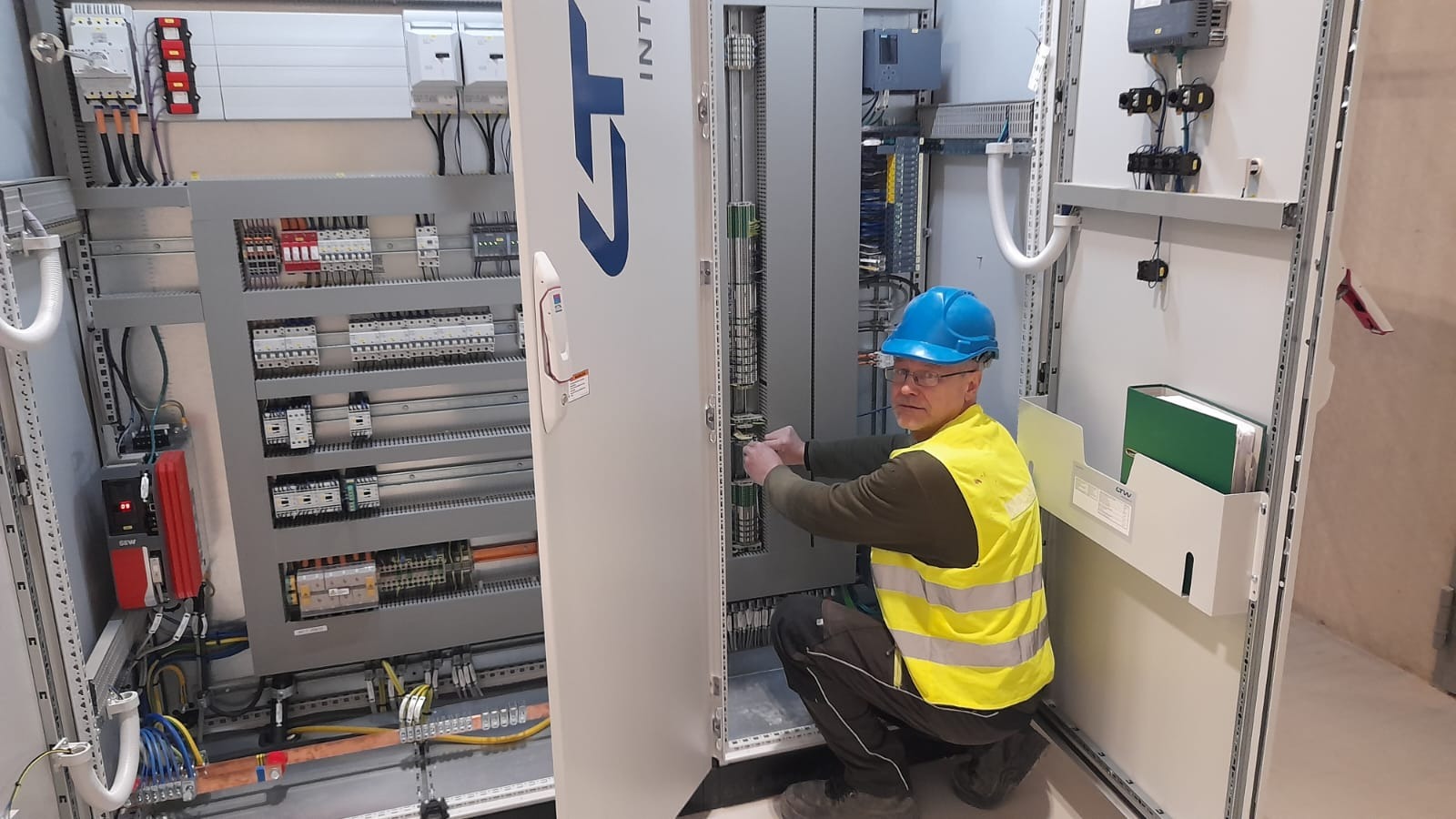 Elektryk w niebieskim kasku i żółtej kamizelce, kuca przed otwartą szafą sterowniczą, podłączając przewody. Widoczna rozbudowana instalacja elektryczna wewnątrz szafy i na ścianie obok.