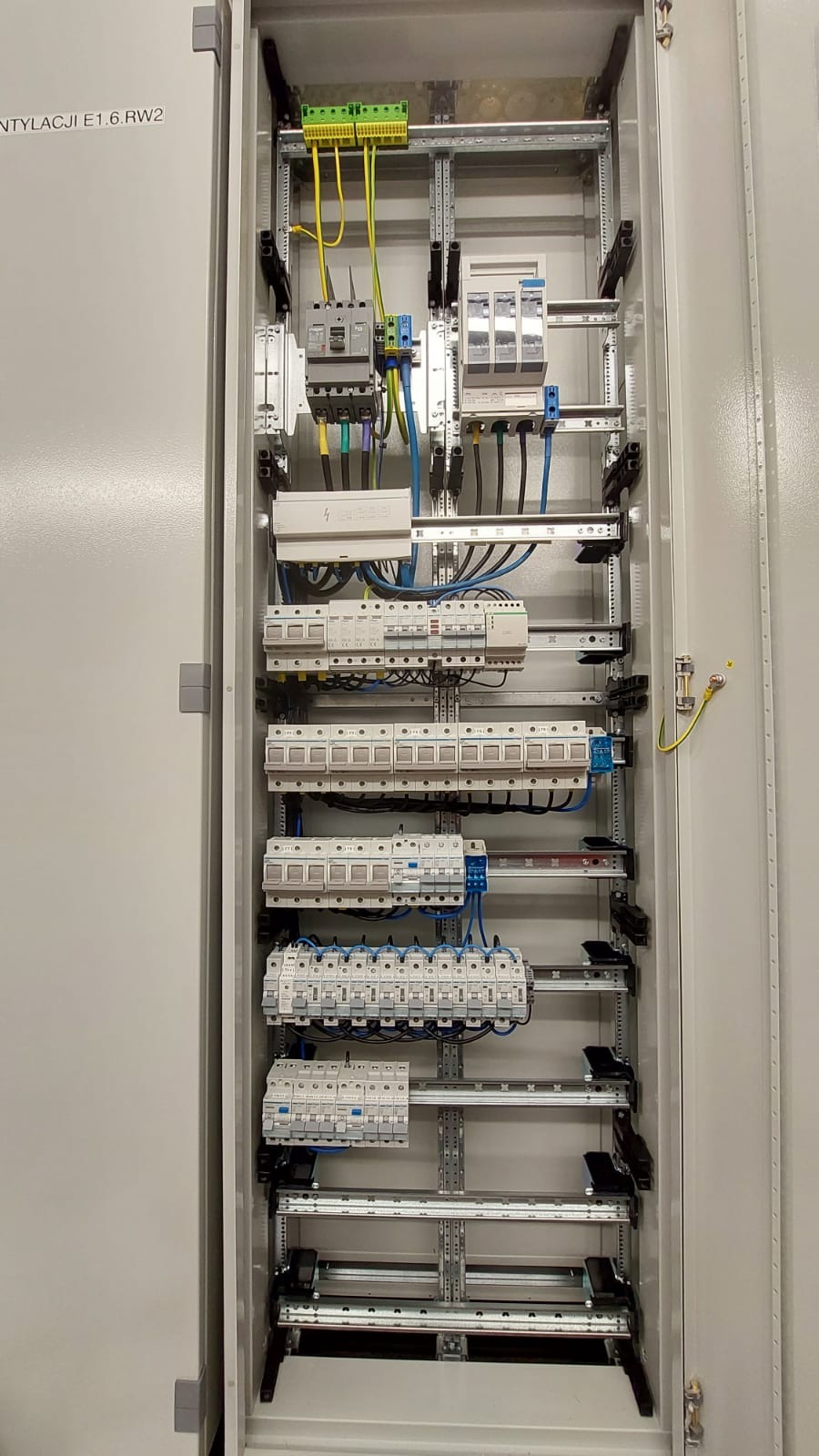 Wnętrze szafy elektrycznej z zamontowanymi bezpiecznikami, wyłącznikami i przewodami, widoczny żółto-zielony przewód uziemiający.
