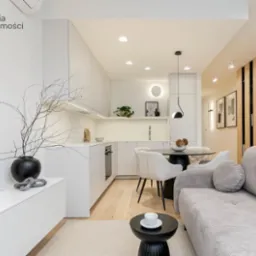 Jasne wnętrze mieszkania z aneksem kuchennym, okrągłym stołem, szarą sofą i dekoracjami w postaci gałęzi w czarnym wazonie.