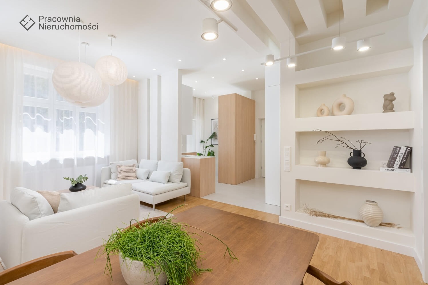 Jasne, przestronne wnętrze mieszkania z białymi sofami, drewnianym stołem z rośliną w doniczce na pierwszym planie, półkami z dekoracjami i oświetleniem punktowym.