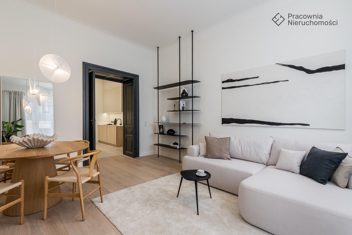 Jasne wnętrze mieszkania z okrągłym stołem, krzesłami z naturalnego drewna, białą sofą narożną i czarnym regałem na tle otwartych drzwi do kuchni.