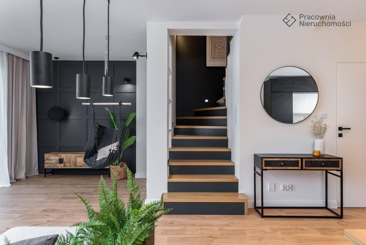 Nowoczesne wnętrze z drewnianymi schodami z czarnymi akcentami, wiszącym fotelem hamakowym obok szafki RTV, okrągłym lustrem nad konsolą i roślinami doniczkowymi.