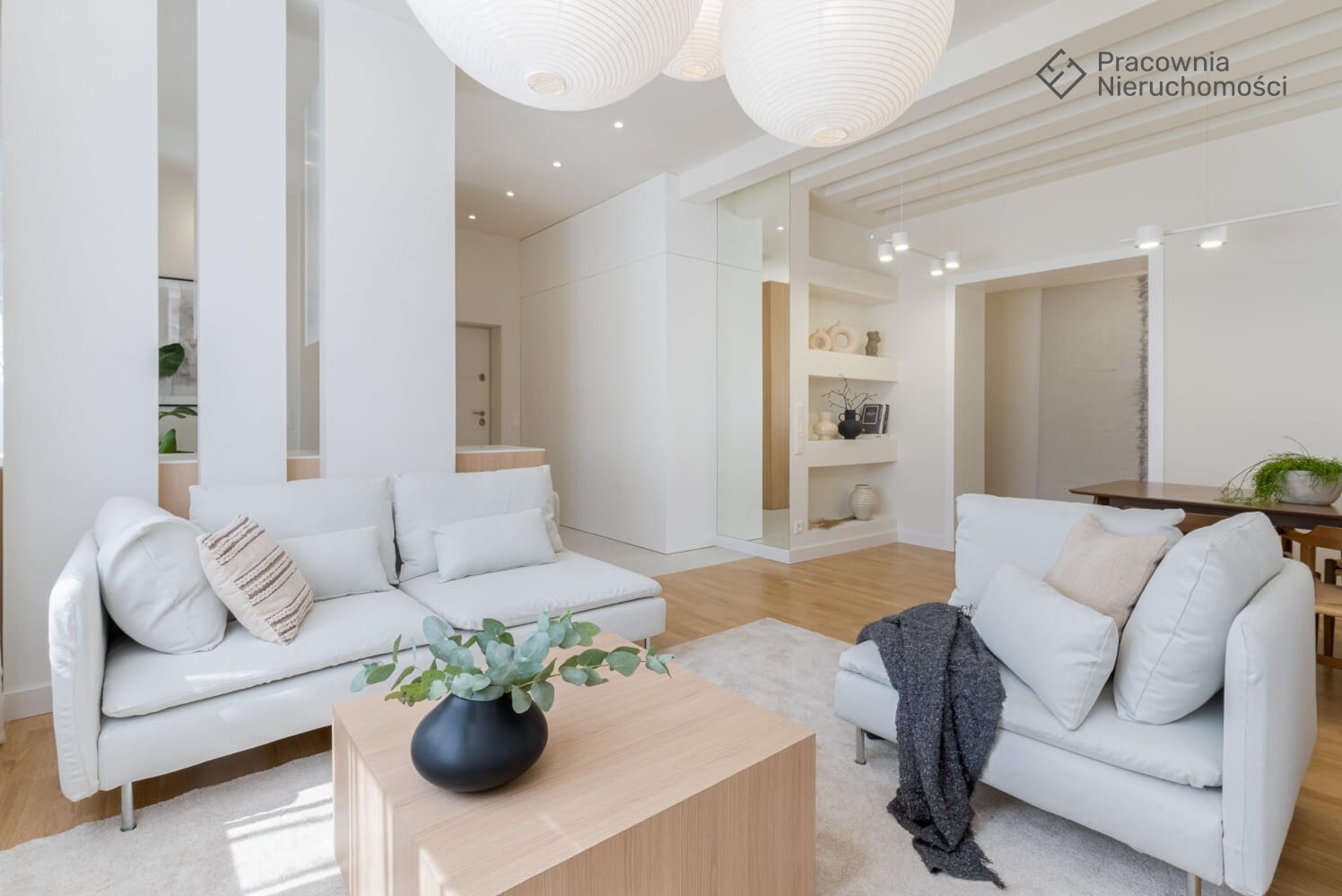 Jasne wnętrze salonu z białymi sofami, drewnianym stolikiem kawowym z czarnym wazonem i zielonymi liśćmi, oraz wiszącymi lampami kulistymi.