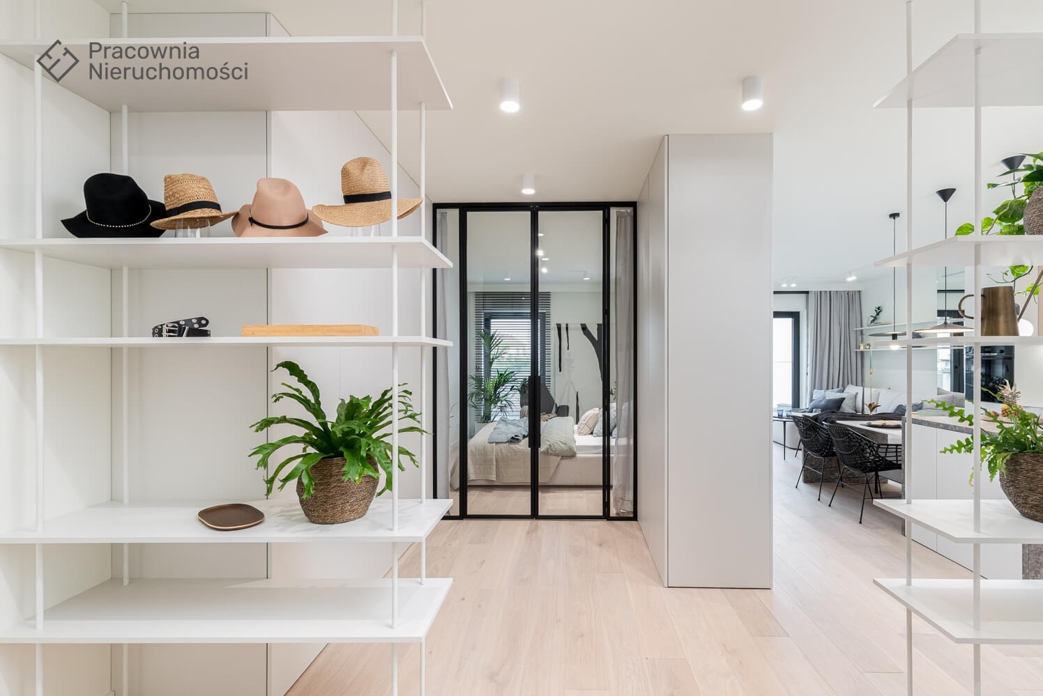 Jasne wnętrze mieszkania z białymi regałami, na których leżą kapelusze, skórzany pasek, drewniana deska i roślina doniczkowa. W tle widoczna sypialnia za szklanymi drzwiami oraz salon z czarnymi...