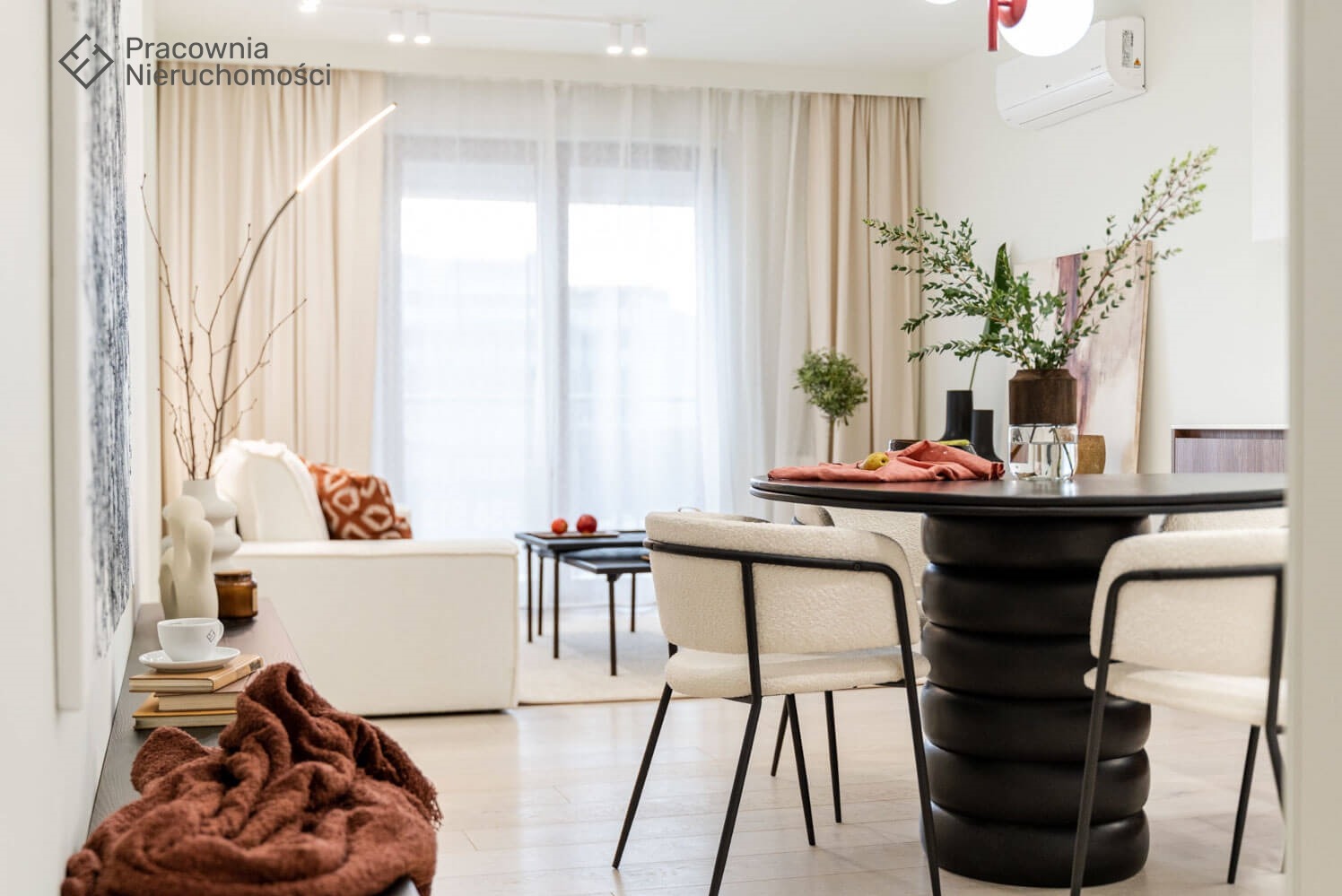 Jasne wnętrze mieszkania z okrągłym, czarnym stołem z podstawą w kształcie ułożonych opon, obitymi krzesłami na metalowych nogach i minimalistycznym salonem w tle.