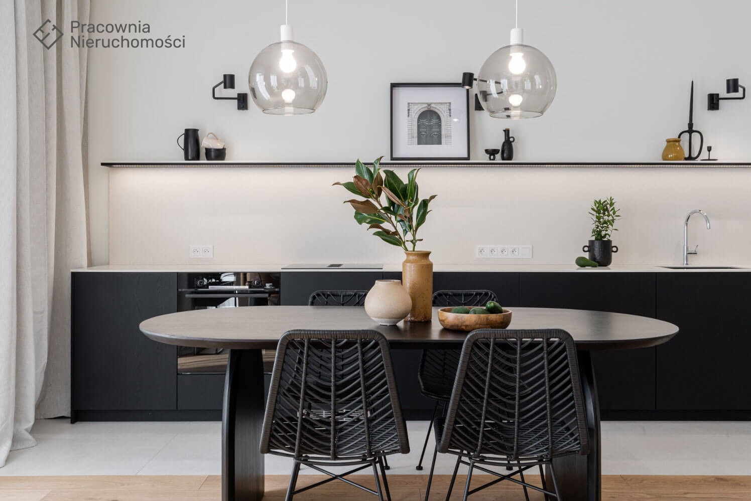 Nowoczesna kuchnia z czarnymi szafkami, blatem w kolorze złamanej bieli, okrągłym stołem z czarnymi krzesłami z rattanu i dekoracyjnymi lampami wiszącymi w kształcie kul.